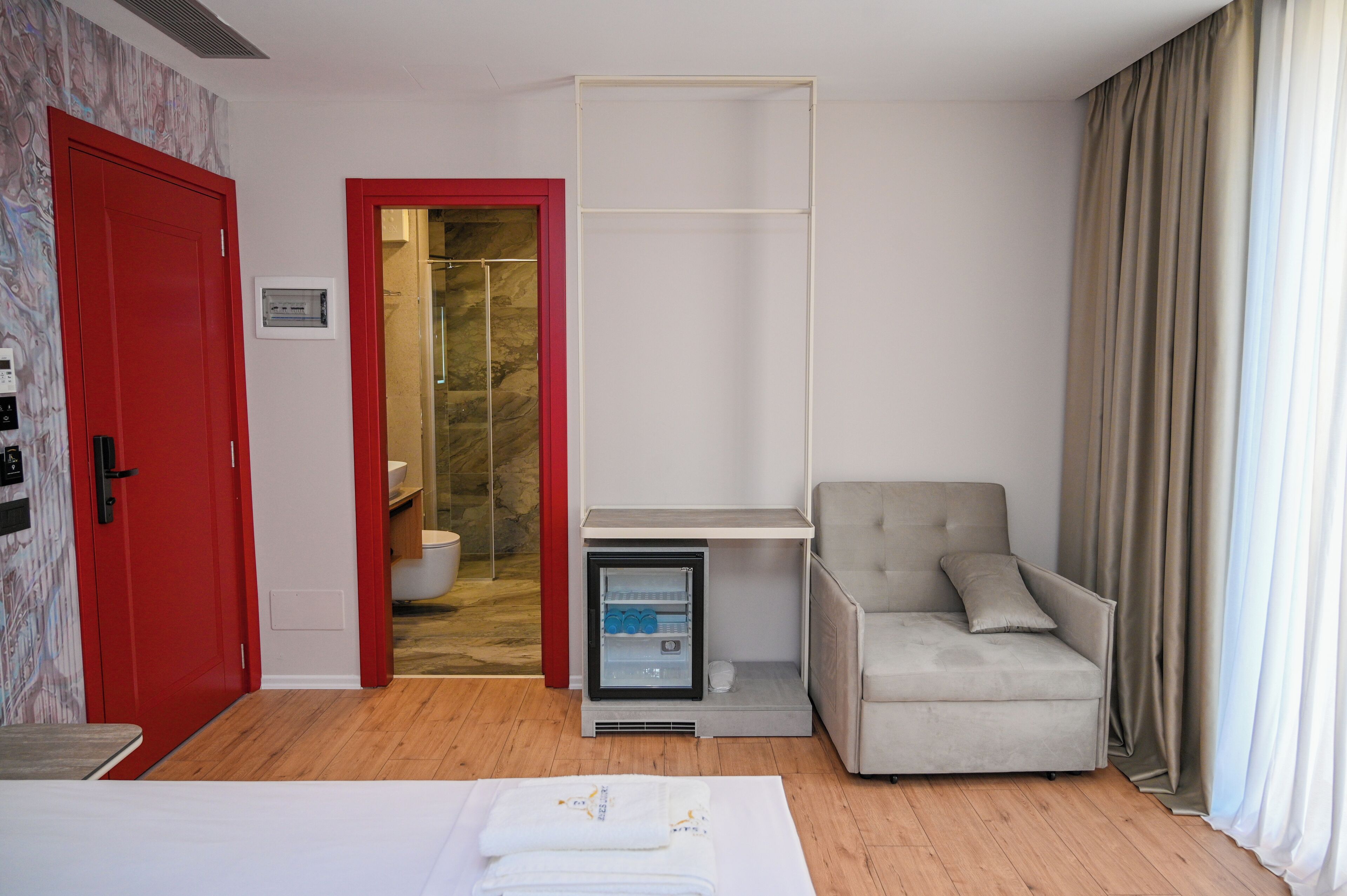 A hotel room with a red door, a mini-fridge, a grey sofa, a bed with white linens, and a bathroom visible through an open door.
