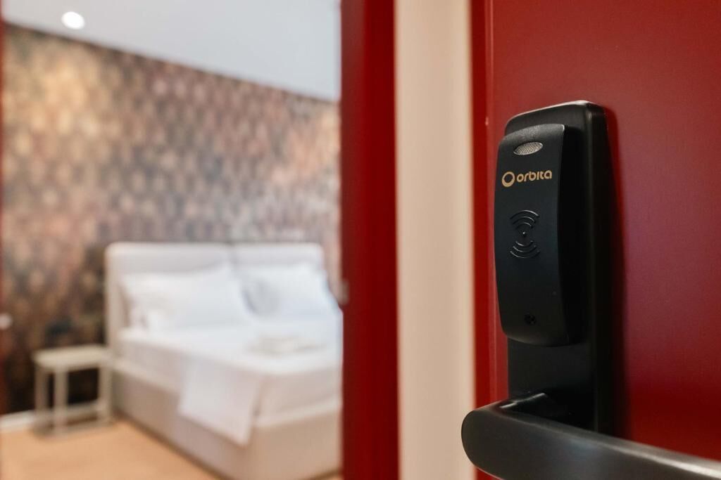 Una habitación de hotel con cama, una mesita de noche pequeña y una puerta roja con timbre.