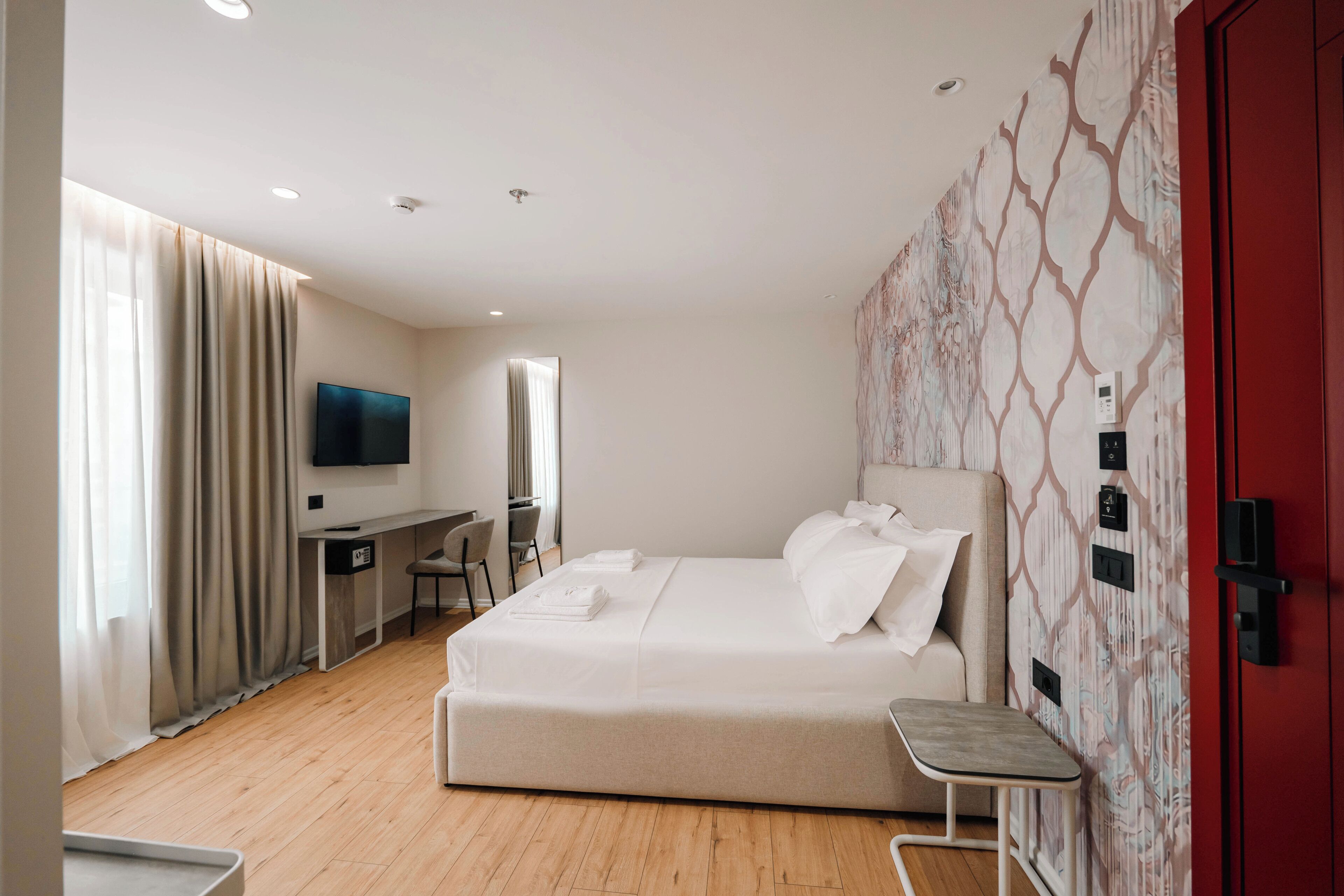 A modern hotel room with a large bed, a desk with a chair, a TV mounted on the wall, and a red door.
