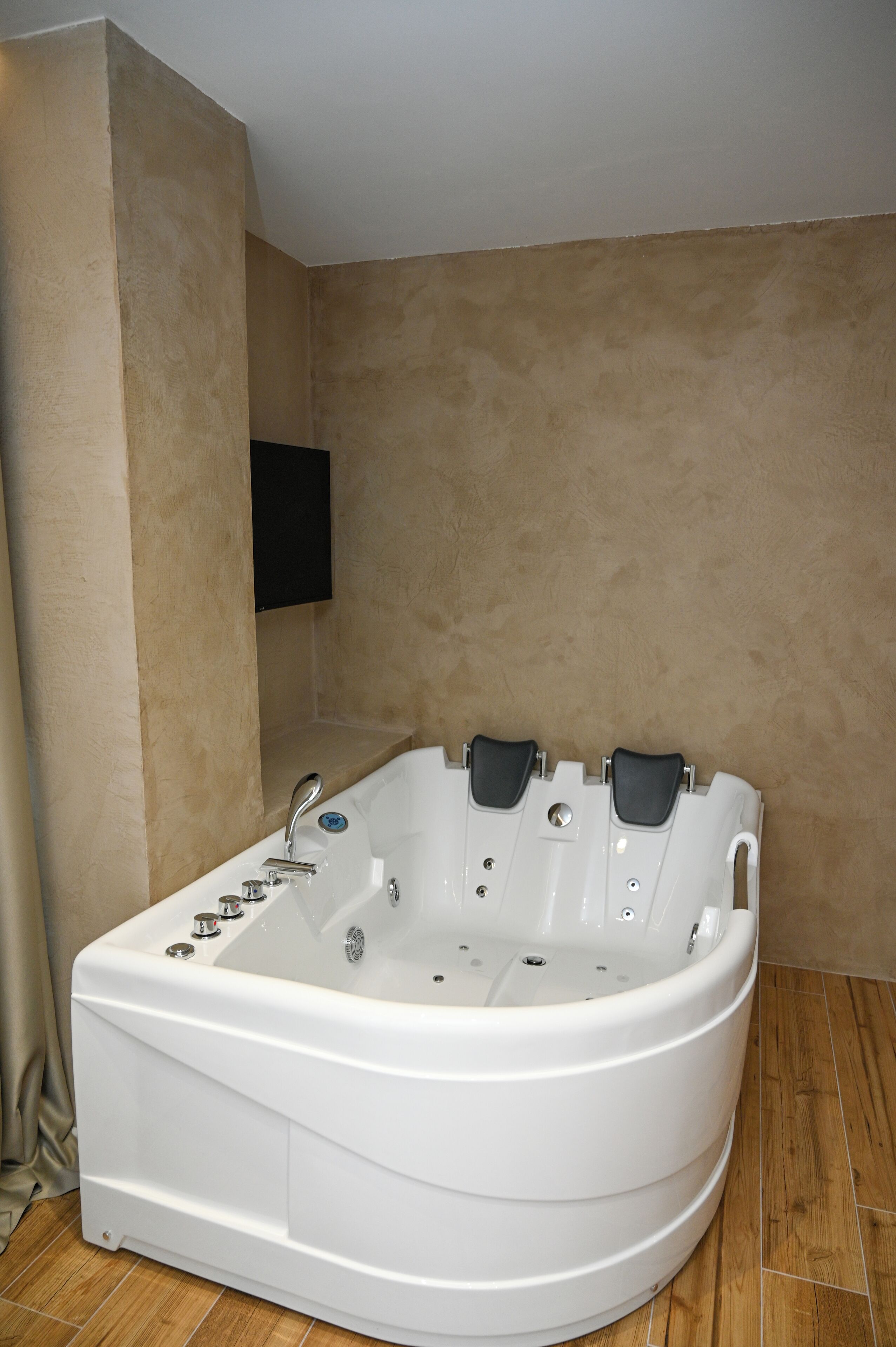A white bathtub with multiple jets in a corner of a room with beige walls and wooden flooring.