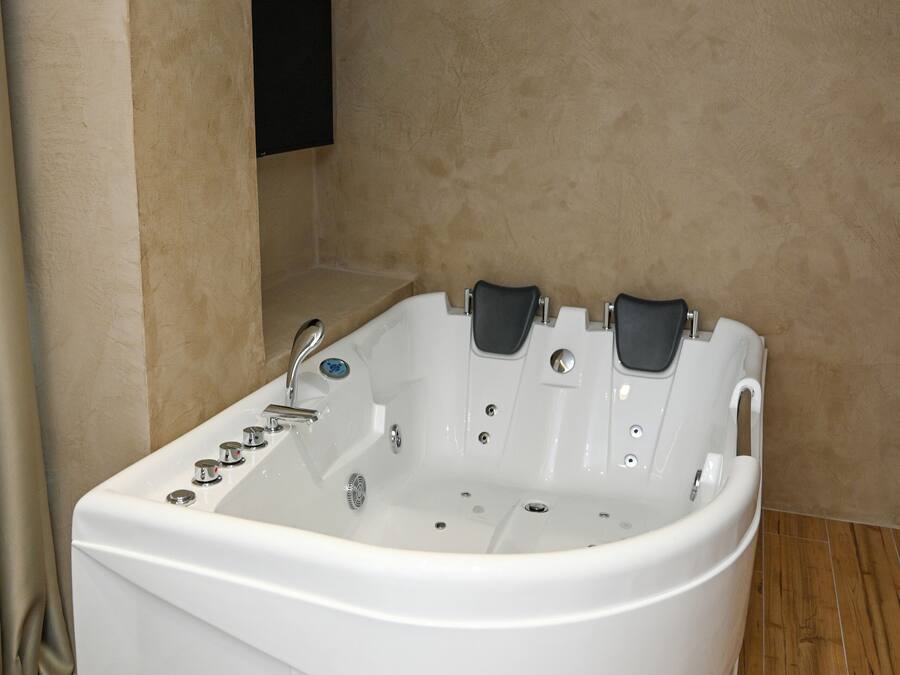 A white bathtub with multiple jets in a corner of a room with beige walls and wooden flooring.