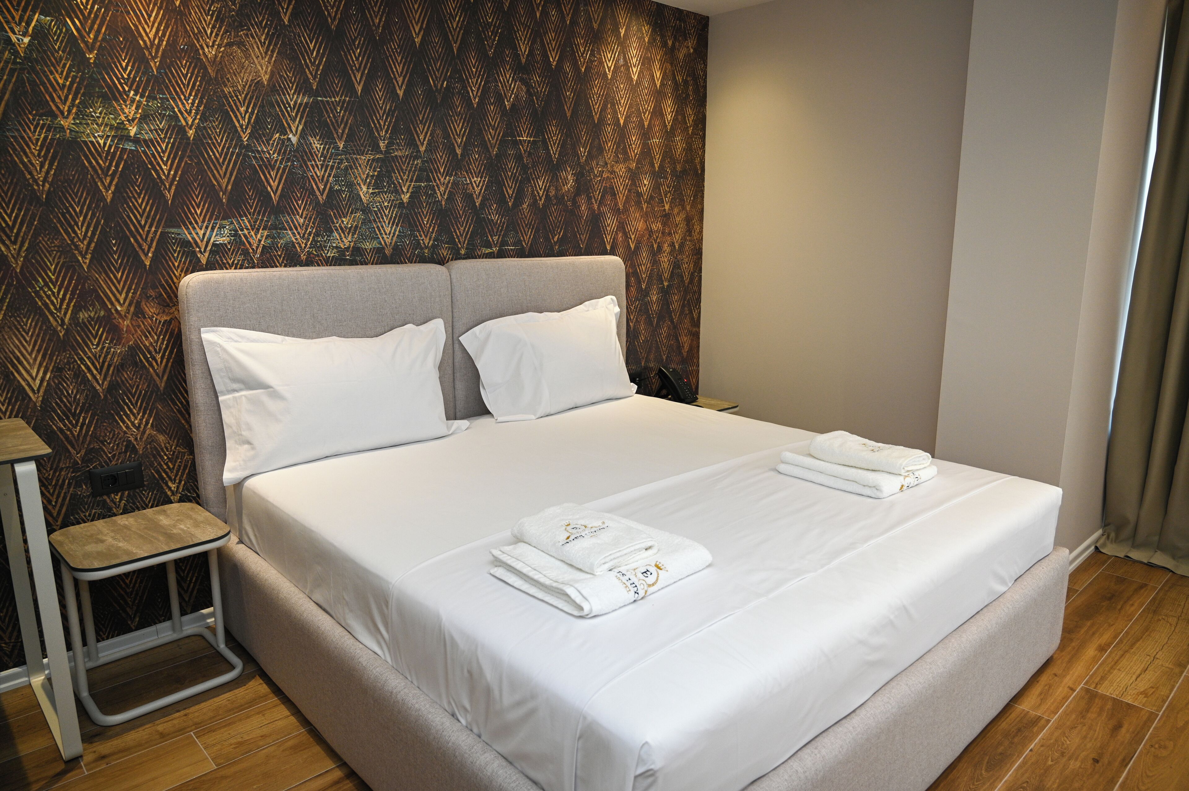 A neatly made bed with white linens and towels, a small wooden stool, and a patterned wallpaper on the wall.