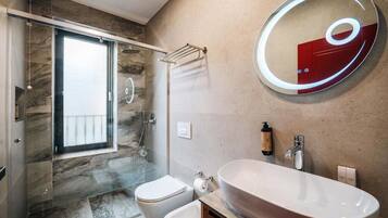 A modern bathroom with a glass-enclosed shower, a freestanding toilet, a large oval sink, and a round mirror with a red accent.