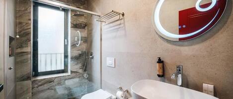 A modern bathroom with a glass-enclosed shower, a freestanding toilet, a large oval sink, and a round mirror with a red accent.