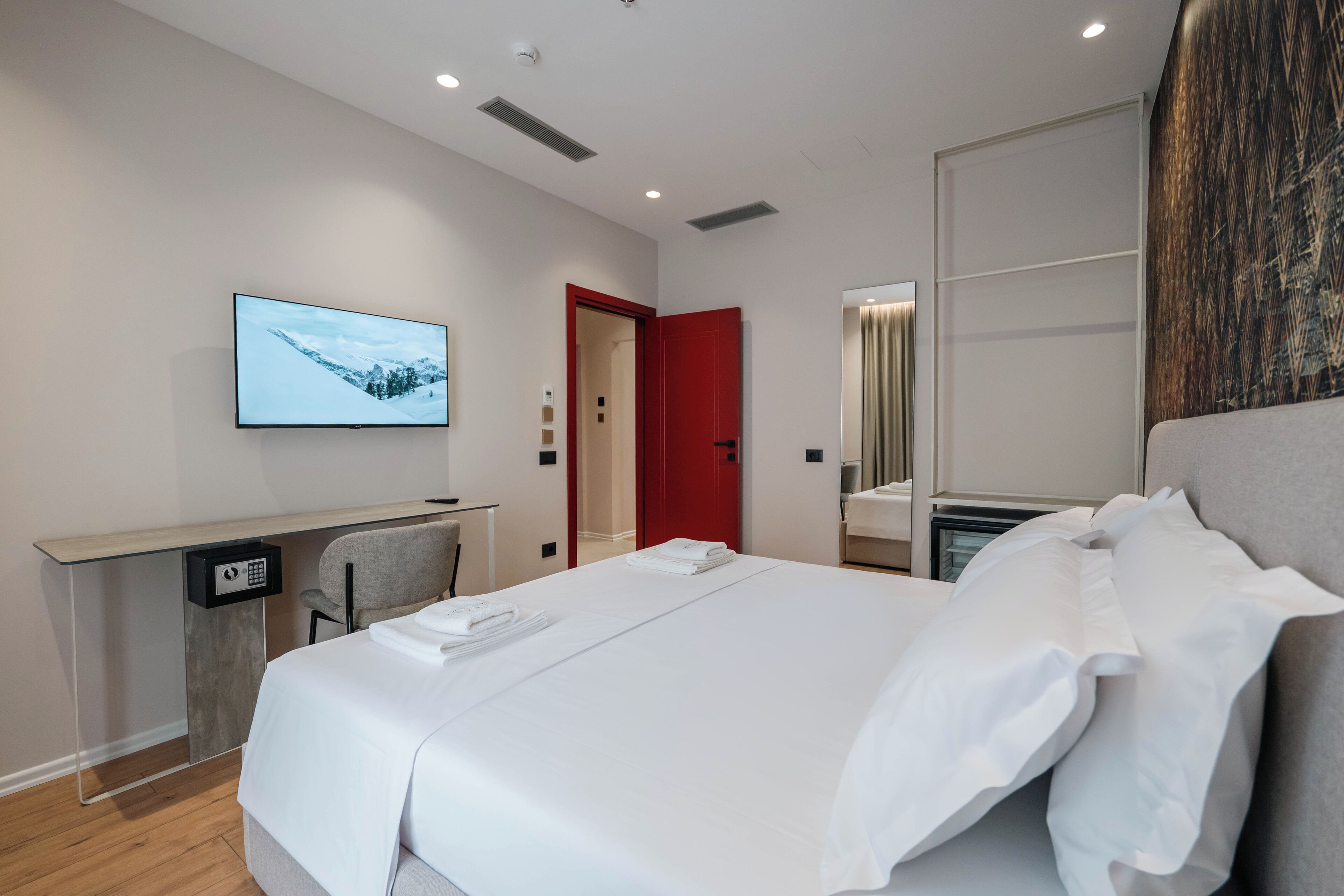 Ein modernes Hotelzimmer mit einem Bett, einem Schreibtisch mit Computer, einem Fernseher und einer roten Tür.