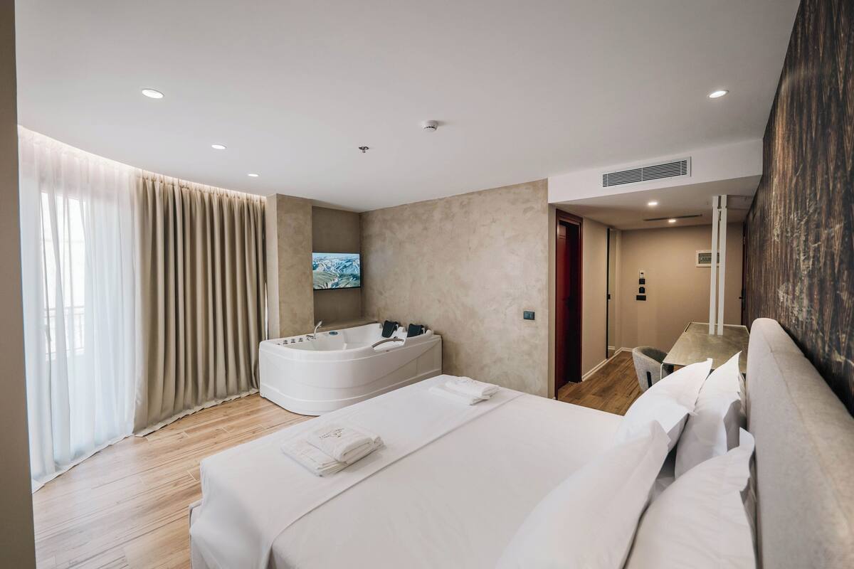 A modern hotel room with a large bed, a jacuzzi, and a TV mounted on the wall.