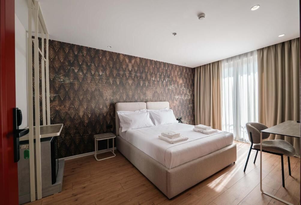 A modern hotel room with a large bed, a small bedside table, a desk with a chair, and a view of the outdoors through large windows.
