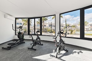 A gym with stationary bikes and a rowing machine, large windows with a view of buildings and palm trees.