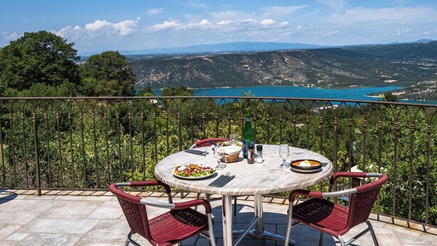 A round table with a white tablecloth, surrounded by red wicker chairs, set on a balcony overlooking a scenic landscape with a lake and mountains. - Camping de l'Aigle (Aiguines)