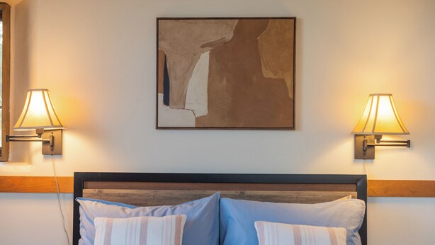 Una cama bien hecha con ropa de cama azul y blanca, dos lámparas de noche y un cuadro abstracto grande en la pared.