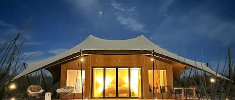 A tent-style building with a large window, a porch with seating, and a sandy ground.