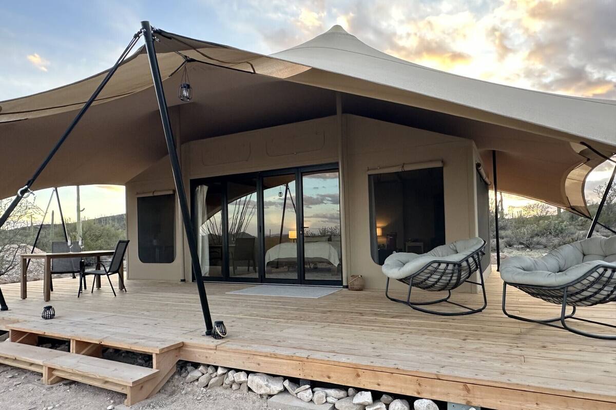 A modern outdoor patio with a wooden deck, a dining table with chairs, and two cushioned chairs under a large canopy.
