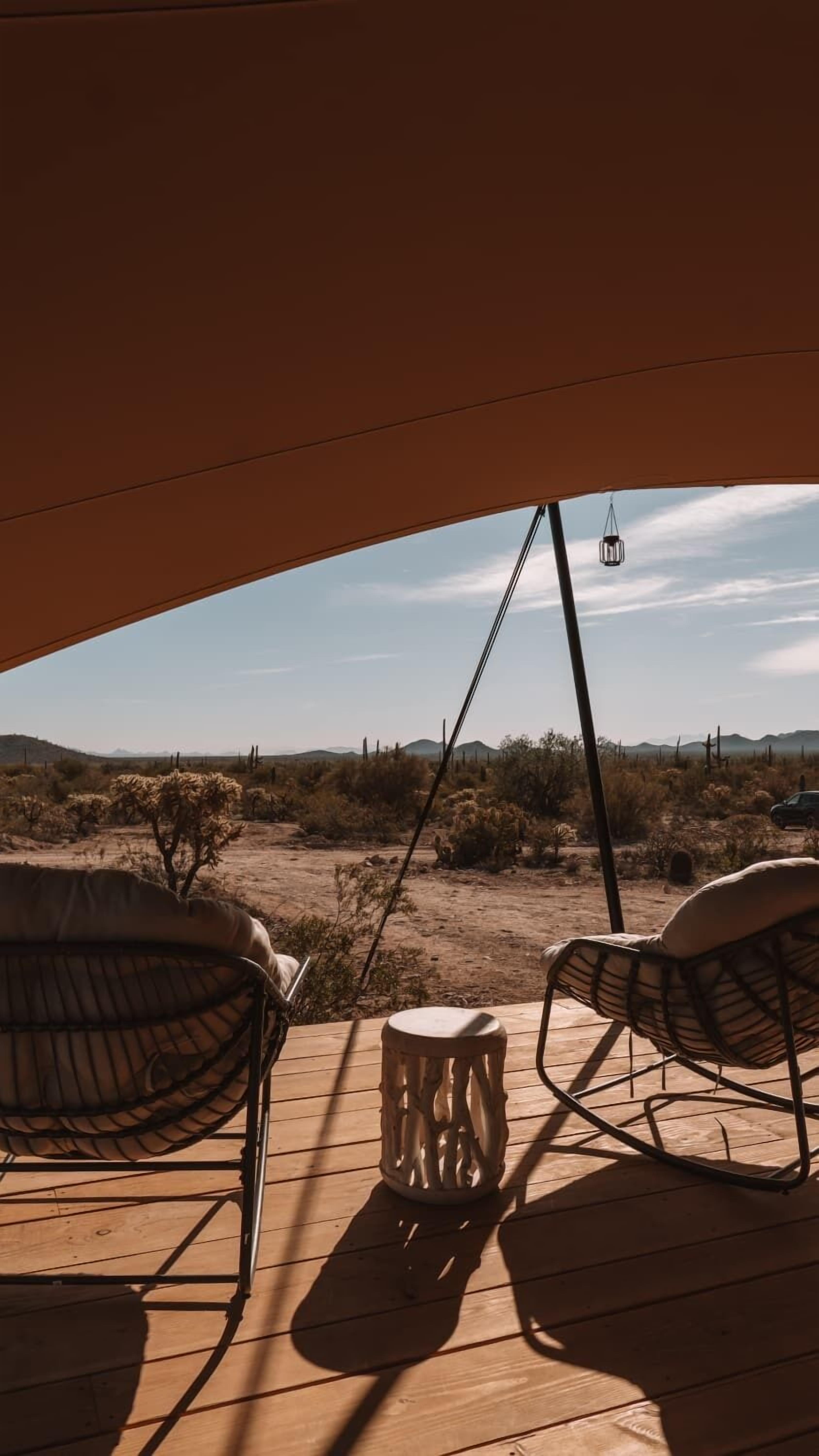 Een houten terras met twee rieten stoelen en een klein rond tafeltje, met uitzicht op een woestijnlandschap met schaarse begroeiing en heuvels in de verte.
