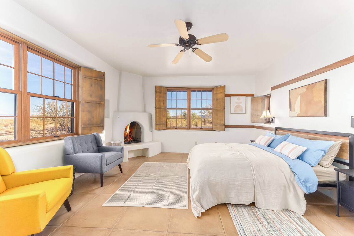 A bedroom with a bed, a desk, a chair, a fireplace, and large windows.