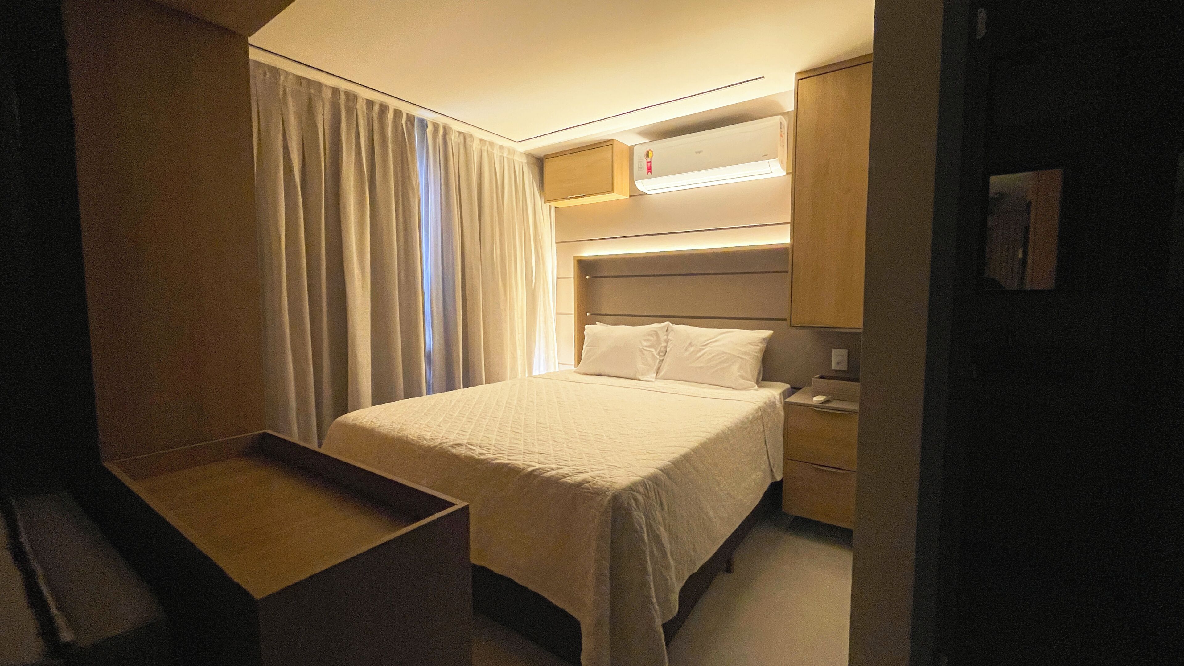 A hotel room with a bed, bedside table, and a built-in air conditioning unit.