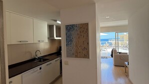 Une cuisine moderne avec des meubles blancs, un plan de travail noir et un plancher en bois. On y trouve un coin repas avec vue sur la mer et un balcon aménagé avec du mobilier d’extérieur.