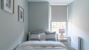 A bedroom with a bed, a nightstand, a lamp, a radiator, and a window with blinds.