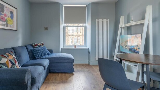 A modern living room with a blue sofa, a wooden floor, a white radiator, a window with blinds, and a TV on a stand.