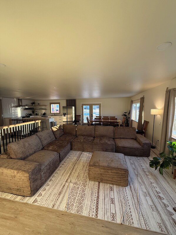 A spacious living room with a large sectional sofa, a patterned rug, and a kitchen in the background. - Enchanting whole home on Nancy lake with WiFi in Willow Alaska! (Willow)