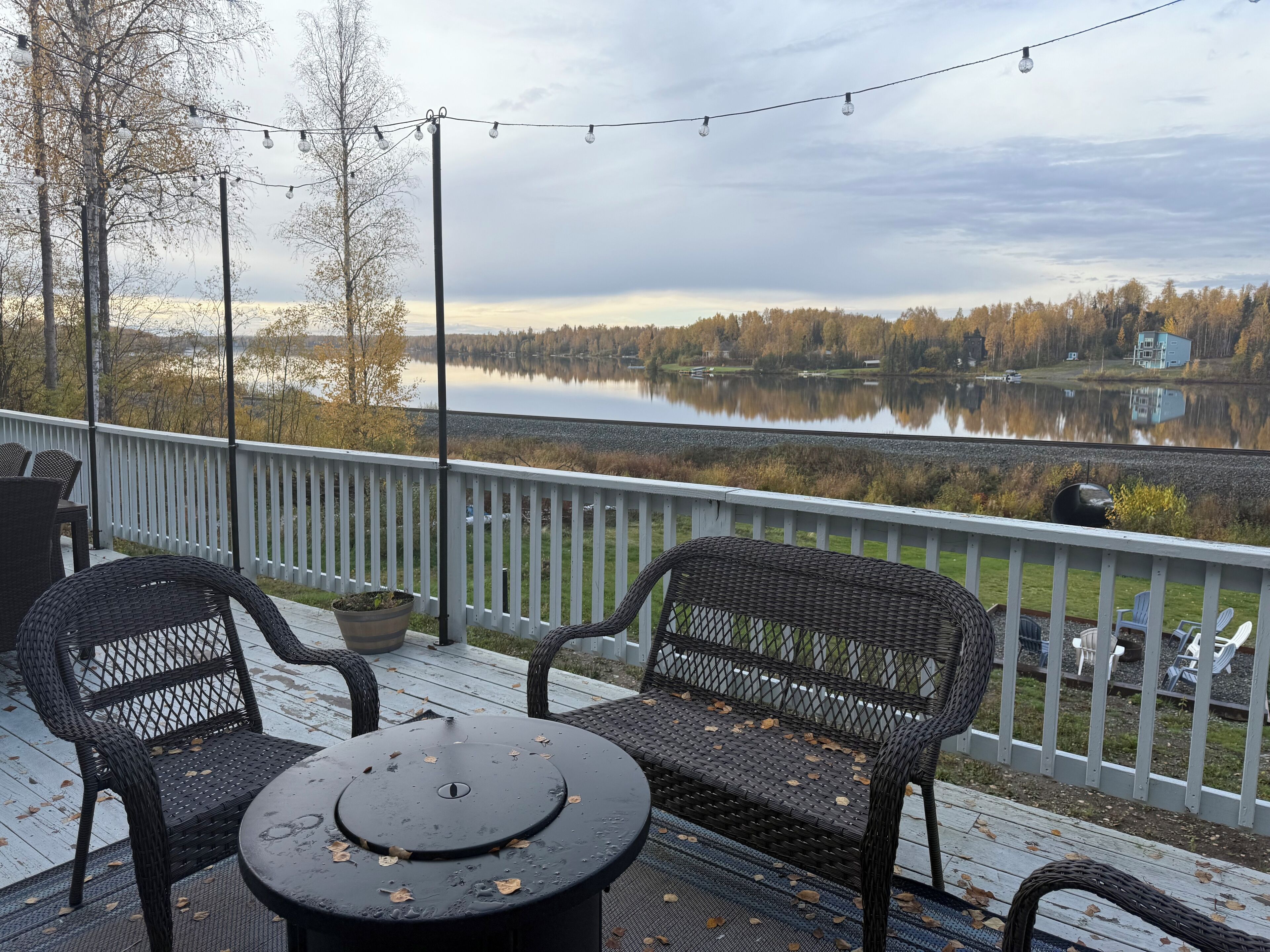 A deck with wicker chairs and a table, overlooking a calm lake with trees and houses in the background.