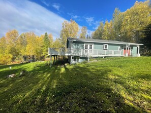 A two-story house with a porch and a deck, surrounded by a grassy yard and trees. - Enchanting whole home on Nancy lake with WiFi in Willow Alaska! (Willow)