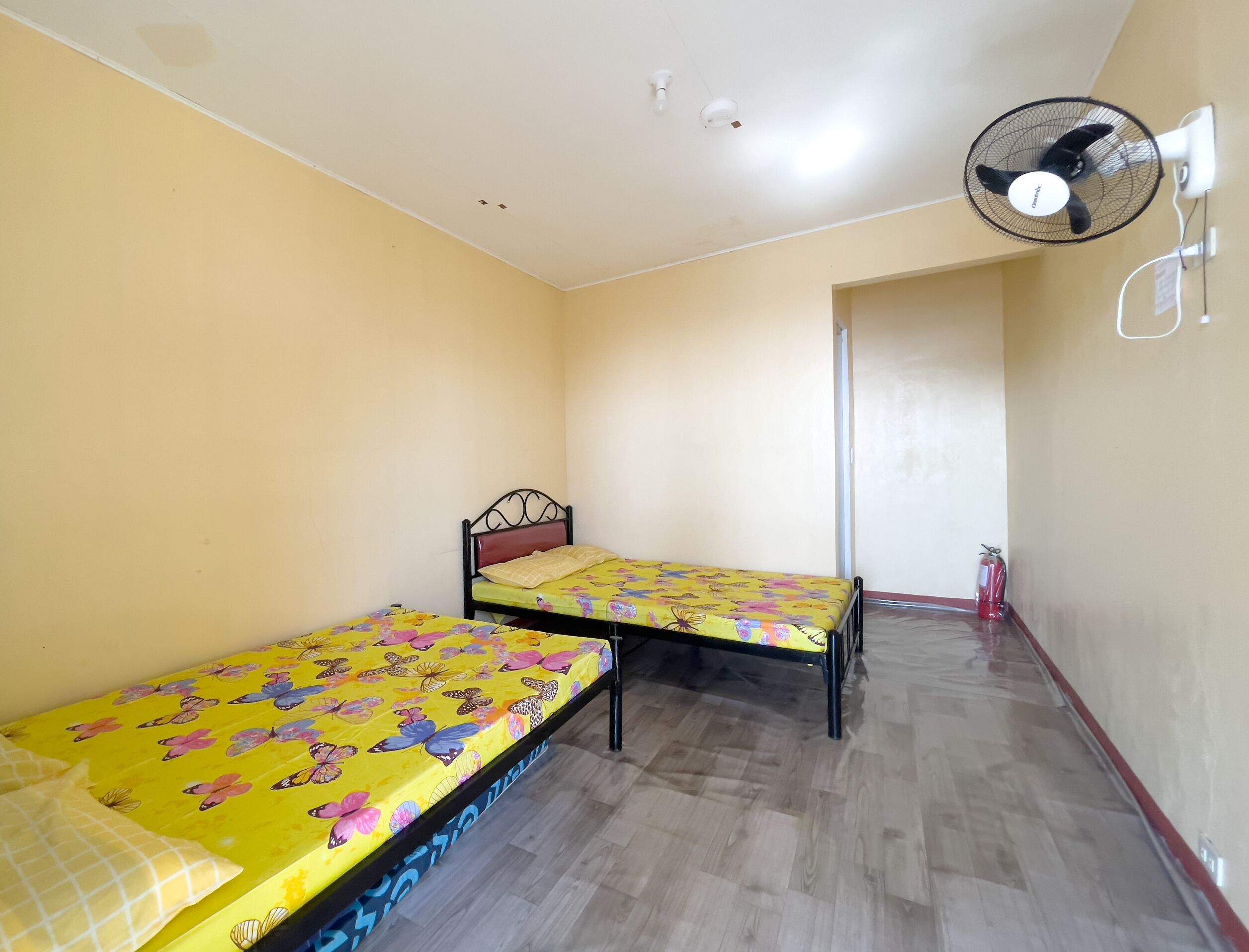 A room with two single beds, a ceiling fan, and a door leading to another room.