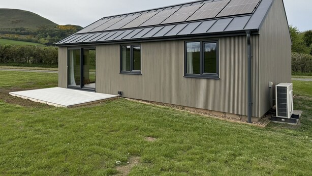 A modern, single-story building with a sloped roof and solar panels. It features a small porch and a large window.