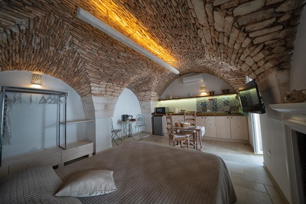 A bedroom with a large bed, a desk, and a kitchenette in an arched stone ceiling room.
