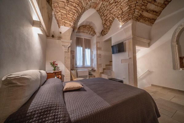 Une chambre avec un plafond voûté en pierre, un lit recouvert d’une courtepointe, une table de chevet avec un vase et une fenêtre avec des rideaux.