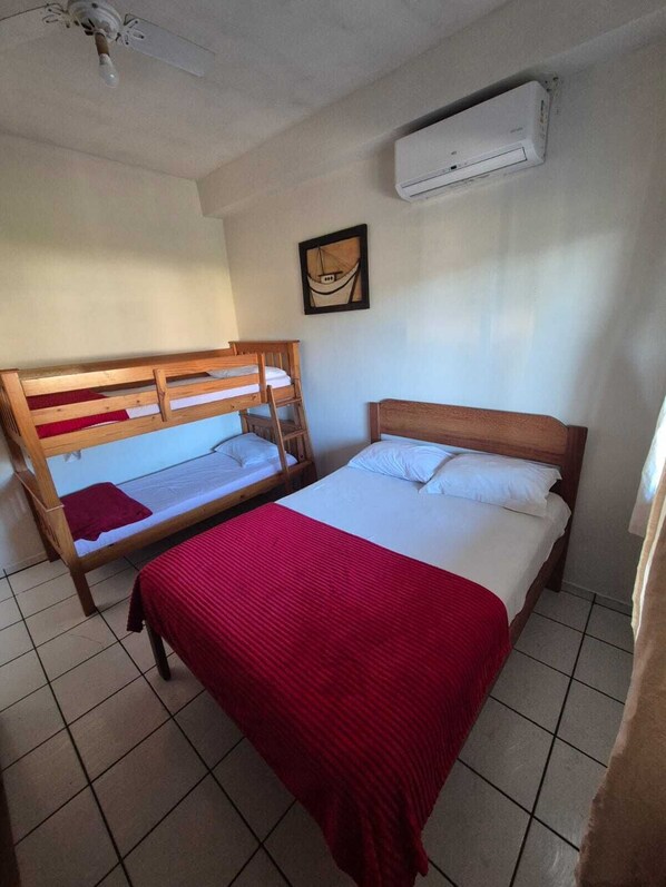 A room with two wooden bunk beds, a single bed, a red blanket, and an air conditioning unit on the wall.