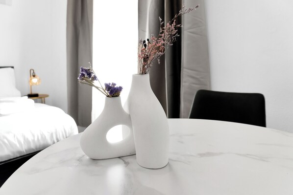 A white marble table with two white vases containing dried flowers. A bed with white linens and a small lamp are visible in the background.