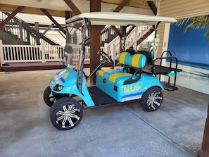 Un carrello da golf blu con dettagli gialli e la scritta 'Beach Life' sul fianco, parcheggiato sotto una tettoia con struttura in legno e un murale sullo sfondo.