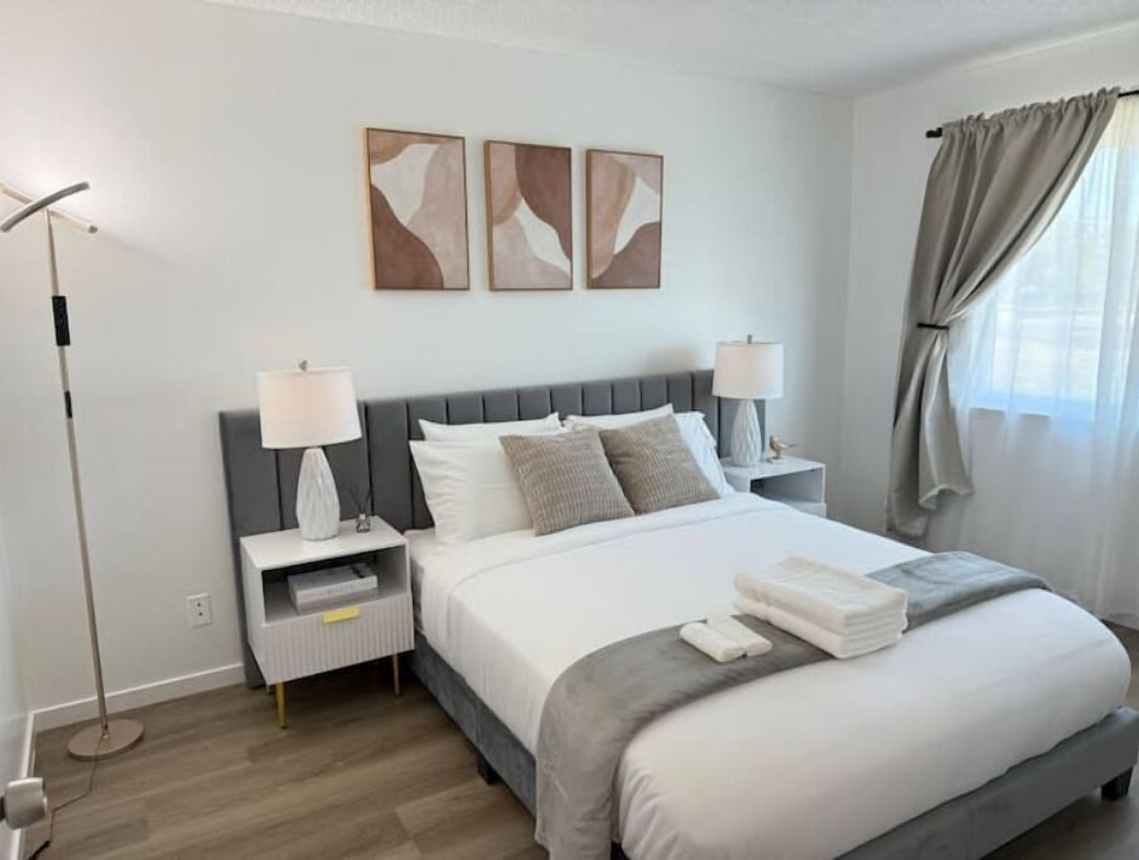 A modern hotel room with a large bed, two bedside tables with lamps, a floor lamp, and abstract wall art.