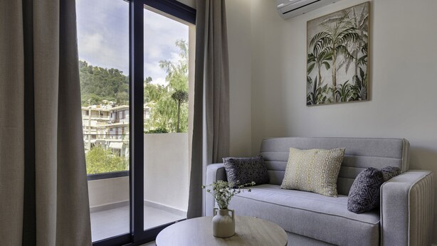 Ein modernes Wohnzimmer mit einem grauen Sofa, einem runden Holztisch und Blick auf eine Wohngegend durch große Fenster.