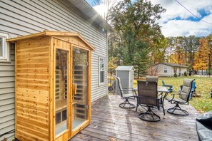 A wooden sauna structure with glass doors, a deck with outdoor furniture, and a house in the background. - Lake George Getaway: Cozy cottage w/ kayaks, cornhole, Xbox, volleyball (Harrison)