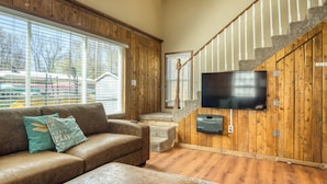 A living room with a brown leather sofa, a television mounted on a wooden wall, and a staircase with white railings. - Lake George Getaway: Cozy cottage w/ kayaks, cornhole, Xbox, volleyball (Harrison)
