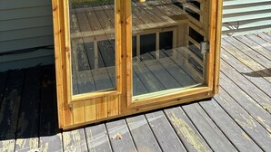 A wooden sauna with a sloped roof and glass doors, situated on a wooden deck. - Lake George Getaway: Cozy cottage w/ kayaks, cornhole, Xbox, volleyball (Harrison)