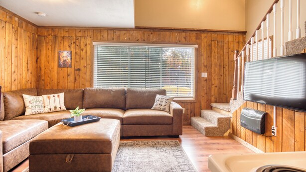 A cozy living room with a brown leather sofa, a matching ottoman, and a flat-screen TV mounted on a wooden wall. - Lake George Getaway: Cozy cottage w/ kayaks, cornhole, Xbox, volleyball (Harrison)