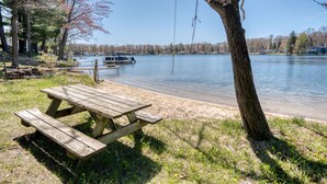 A wooden picnic table by a lakeside with a boat docked in the background. - Lake George Getaway: Cozy cottage w/ kayaks, cornhole, Xbox, volleyball (Harrison)