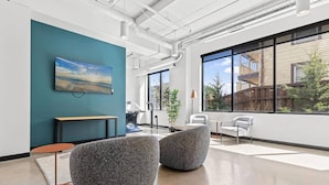 Un espacio de oficina moderno con una pared de acento color aguamarina, un gran ventanal con vistas a edificios y un cuadro de un paisaje.