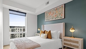 Un dormitorio moderno con una cama grande, dos mesitas de noche, una lámpara y una ventana con vistas, además de un elemento decorativo en la pared.