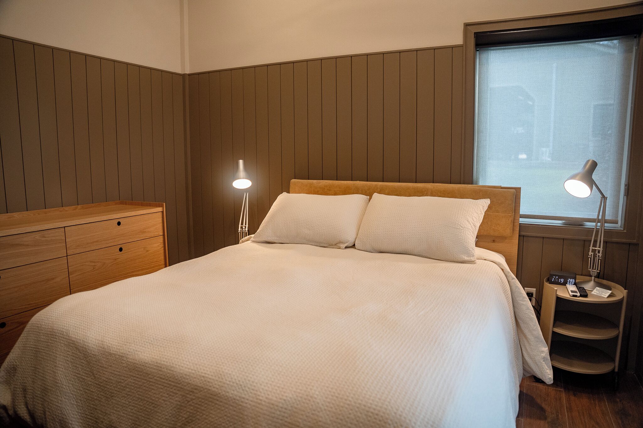 Une chambre à coucher bien rangée, avec une tête de lit en bois, une table de chevet avec une lampe et une fenêtre donnant sur un beau paysage.