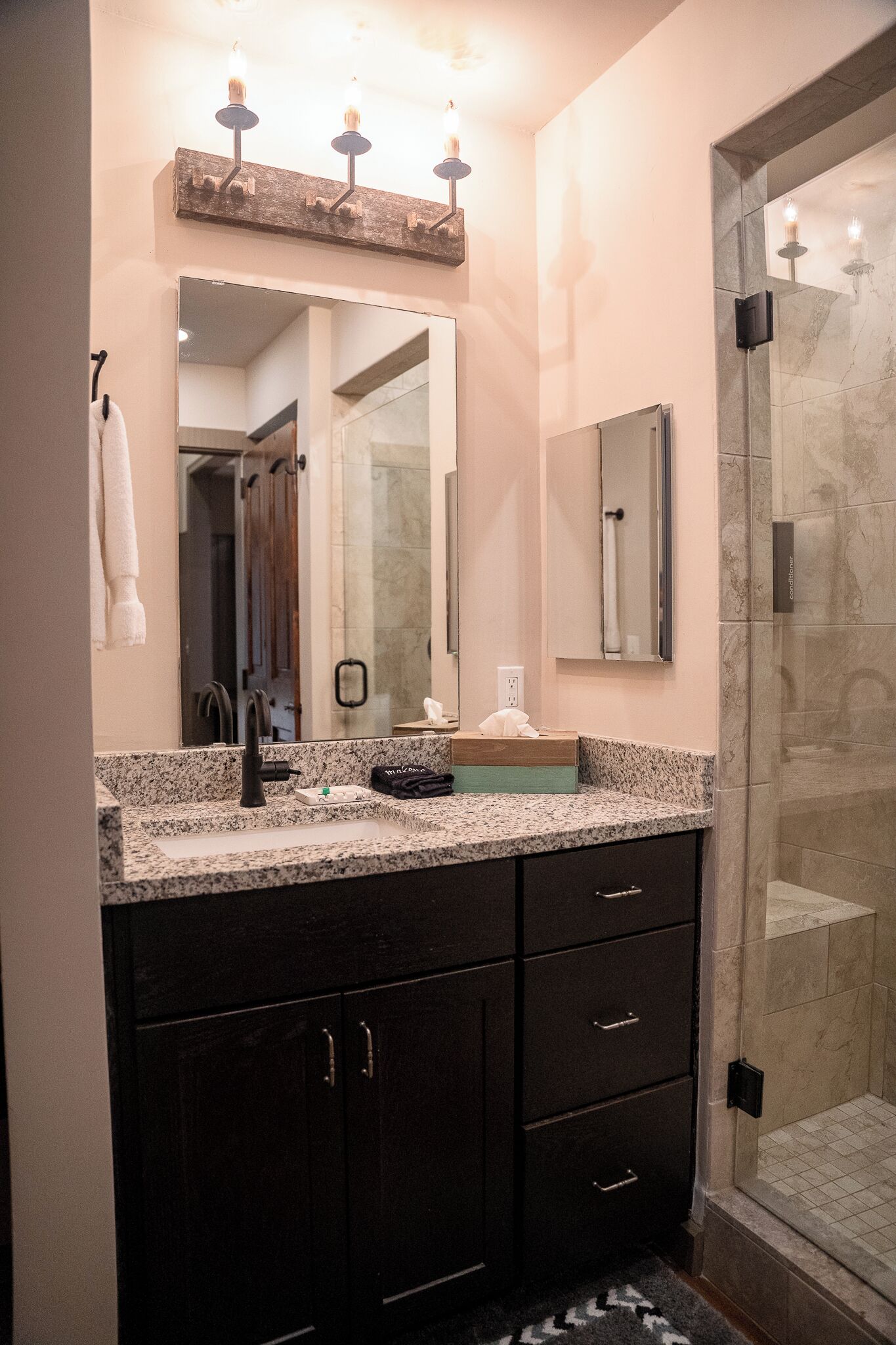 A bathroom with a granite countertop, dark cabinets, a large mirror, and a glass shower enclosure.