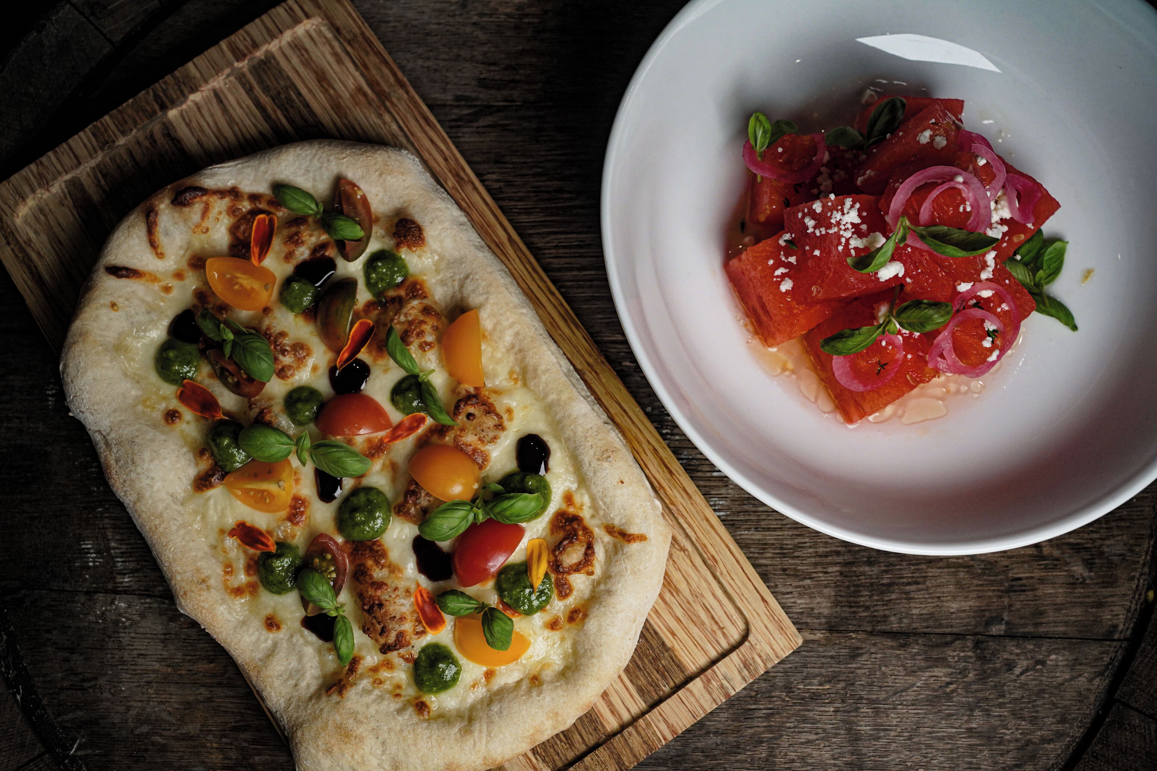 Una pizza con diferentes ingredientes sobre una tabla de madera y un tazón de ensalada con tomates y albahaca.
