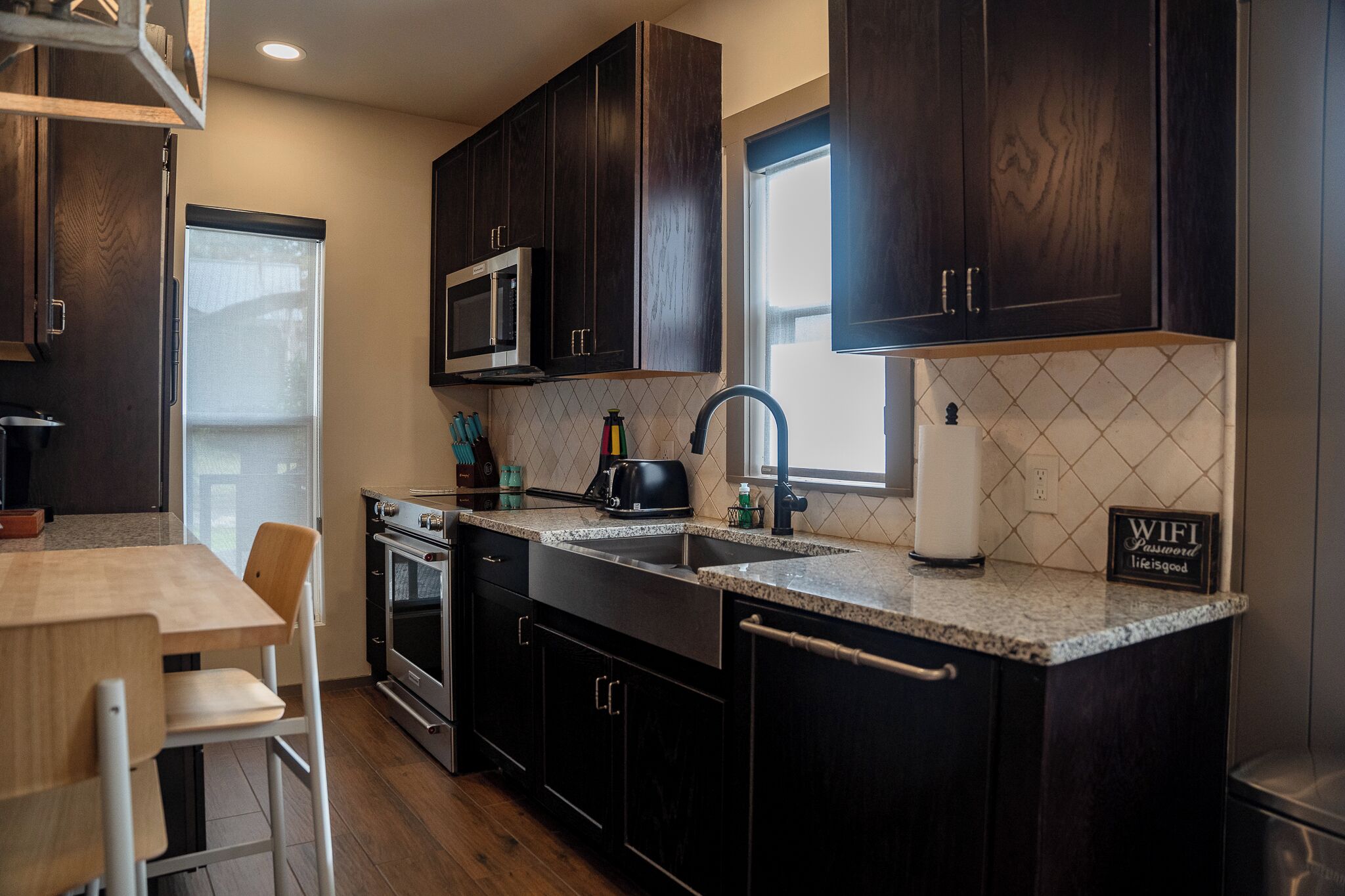 A modern kitchen with dark wood cabinets, a granite countertop, a built-in microwave, a stainless steel sink, and a window providing natural light.