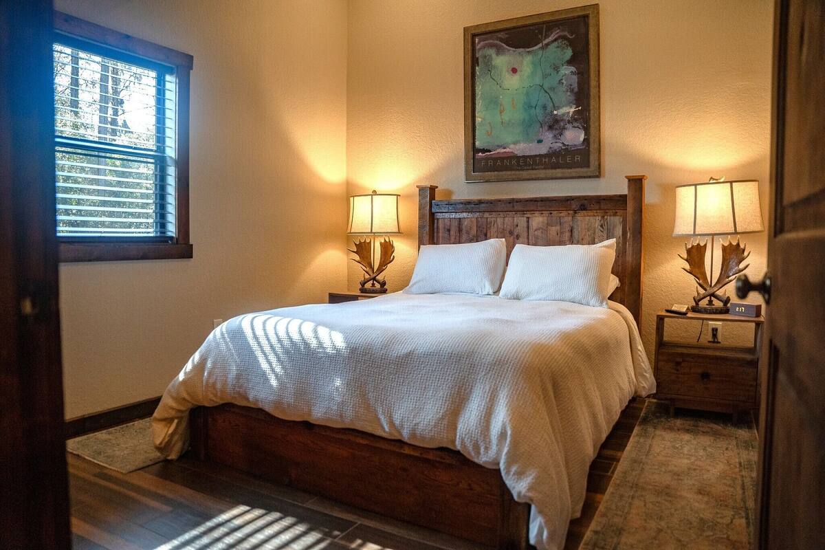 A bedroom with a wooden bed, two bedside lamps, a framed map on the wall, and a window with blinds.