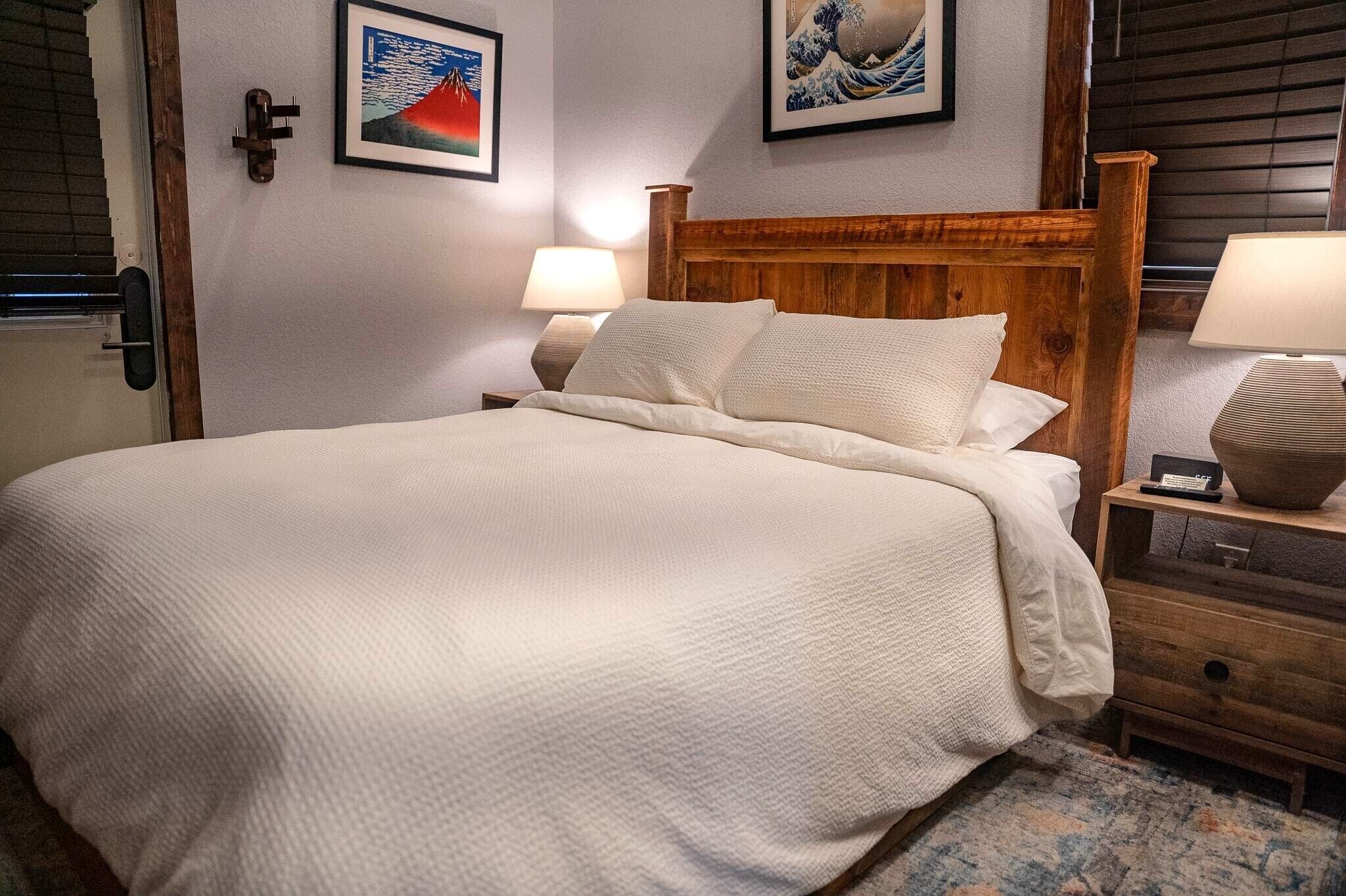 Een net opgemaakt bed met een houten hoofdbord, twee nachtkastjes met lampen, en twee ingelijste kunstwerken aan de muur.