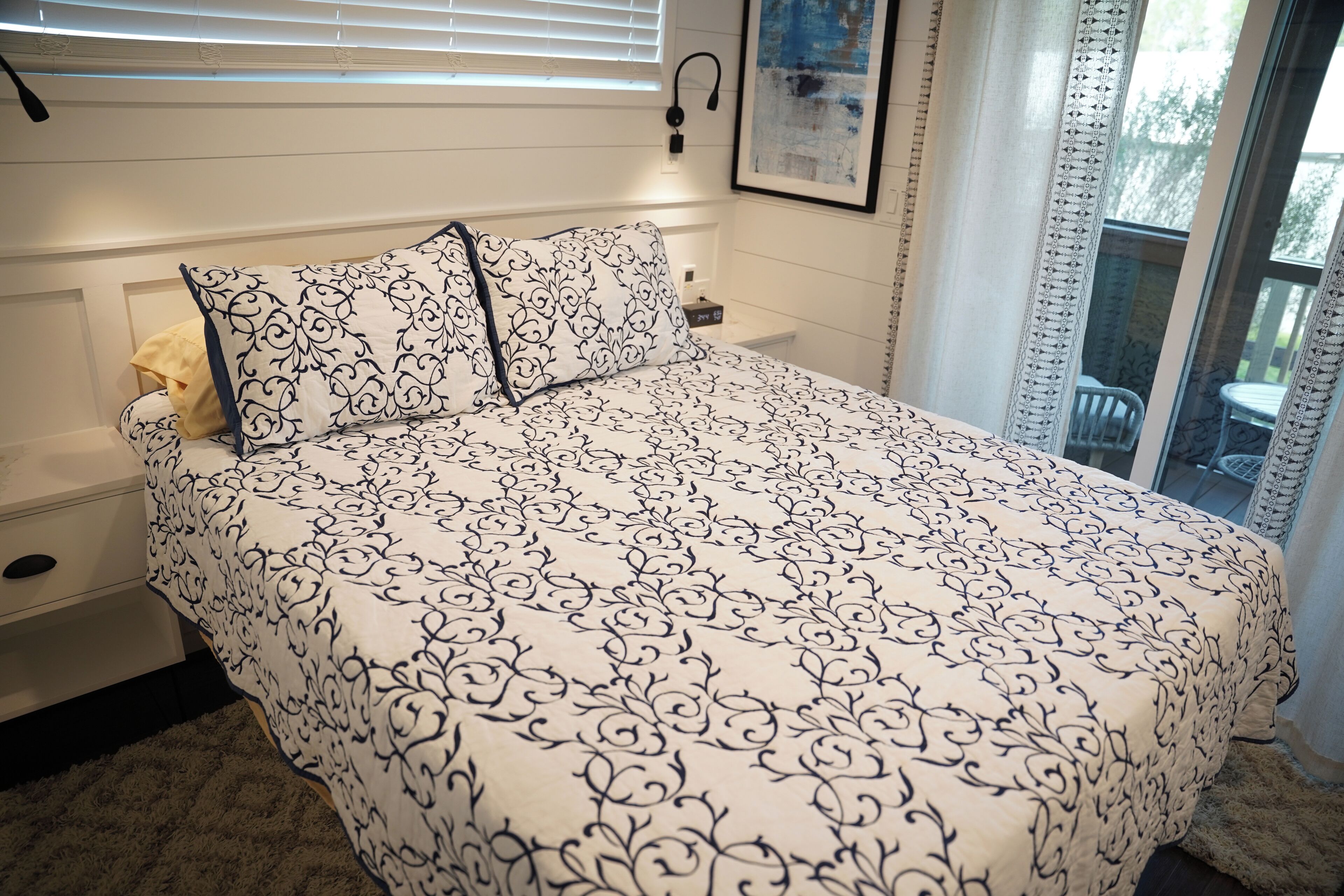 A bedroom with a large bed, a nightstand, a lamp, and a window with blinds.