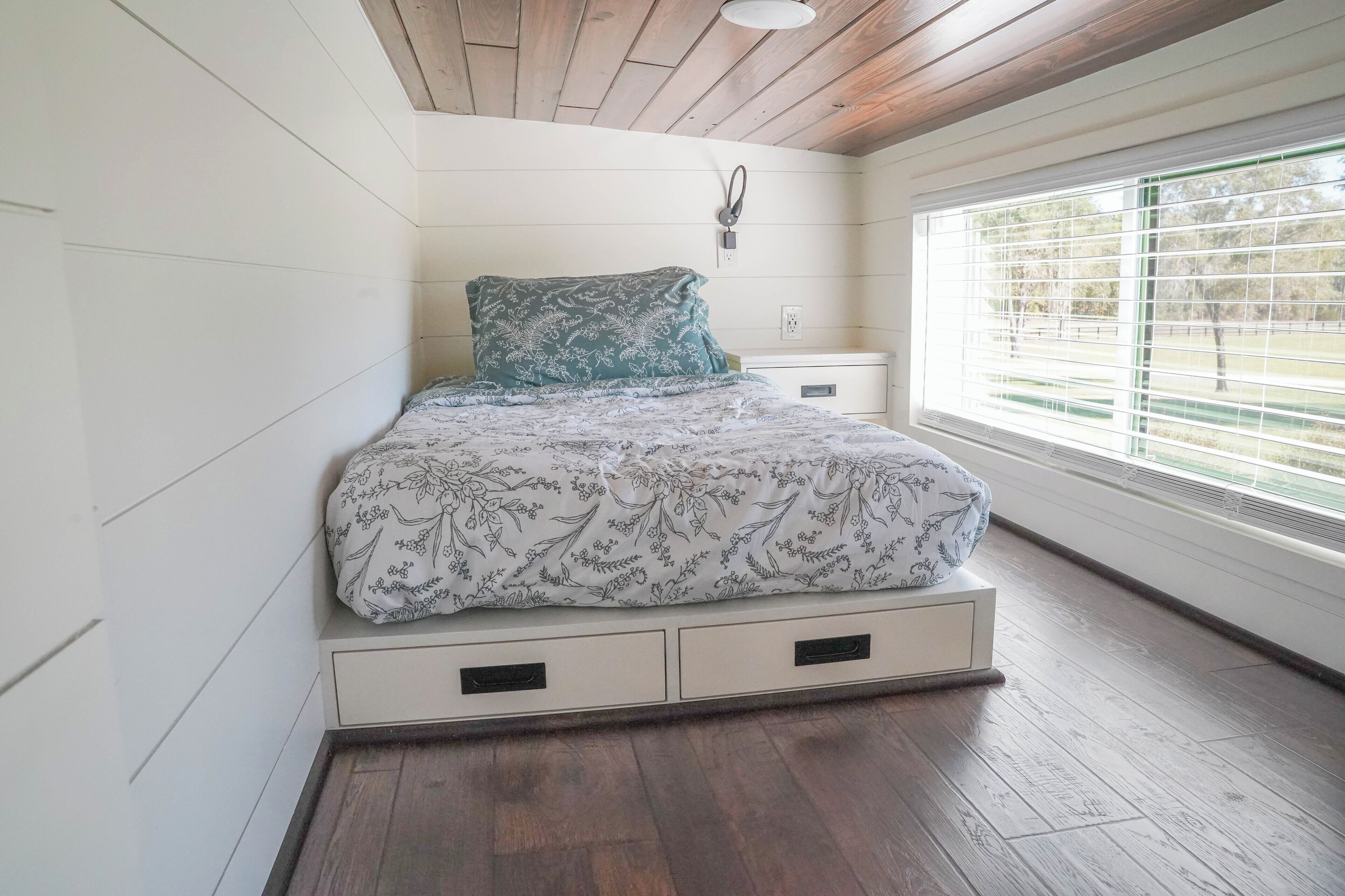 A bedroom with a bed, a nightstand, a wall-mounted lamp, and a window with blinds.
