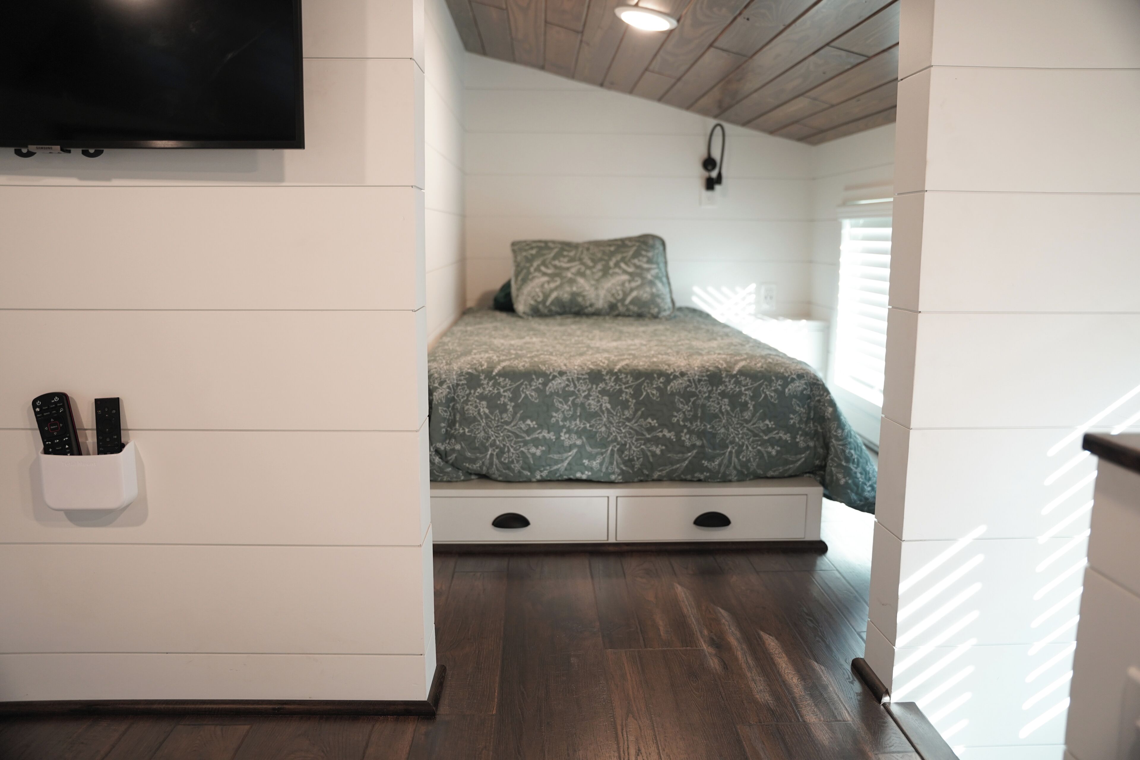 A bedroom with a bed, a TV mounted on the wall, a remote holder, and a window with blinds.